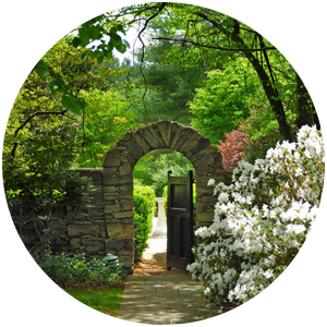 A stone arch with a gate and a green garden surrounding it. White azaleas are in the foreground.