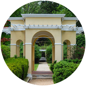 A yellow stucco garden arched gazebo with bushes, trees, and gravel paths